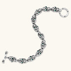 Silver stainless steel skull link bracelet with green crystal eye sockets and engraved detailing, toggle clasp closure.