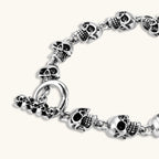 Silver stainless steel bracelet with linked 3D skull charms and a spring-ring clasp, oxidized black detailing, 225mm length.